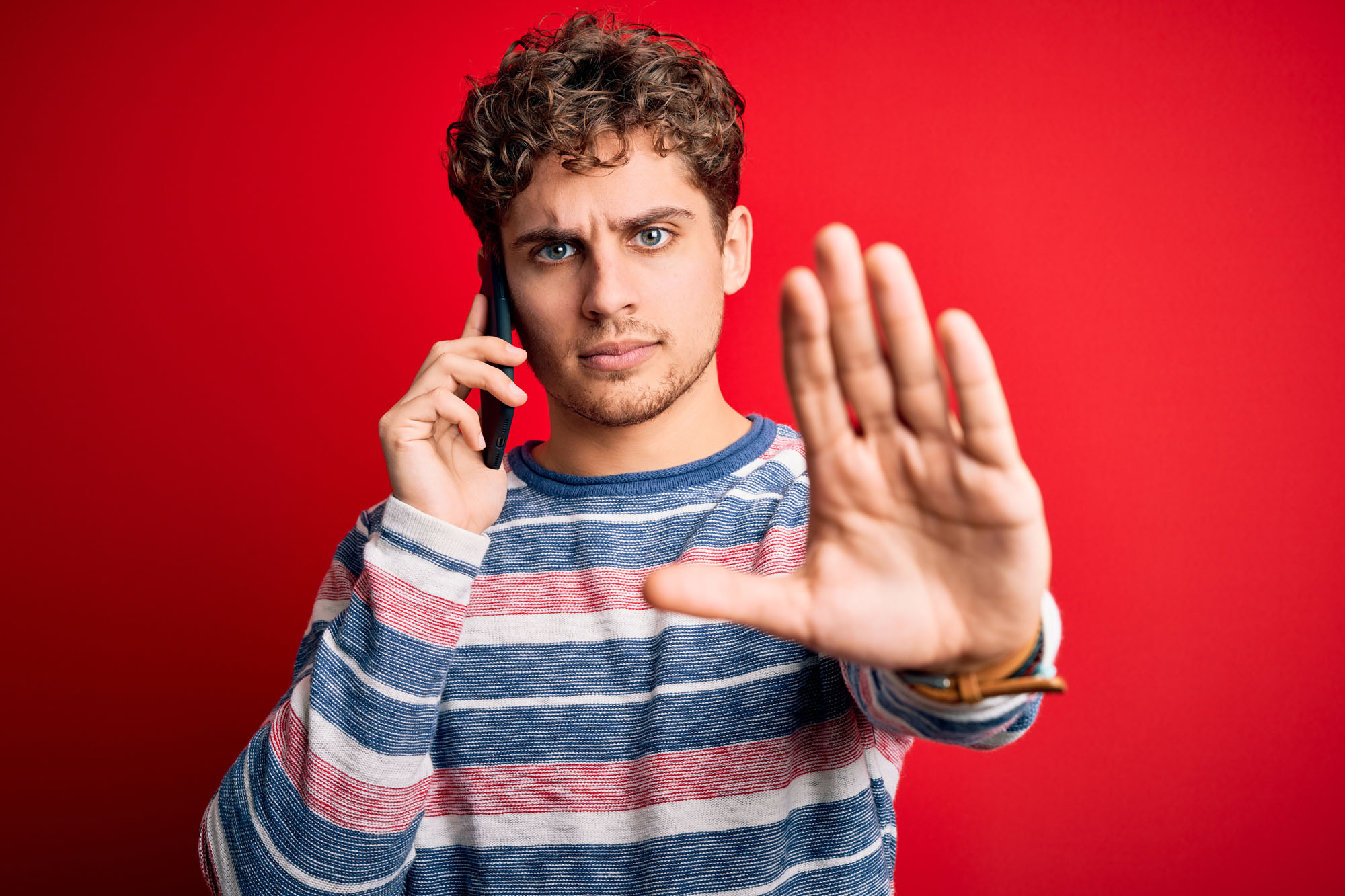 Young blond man with curly hair having conversation talking by the smartphone with open hand doing stop sign with serious and confident expression, defense gesture